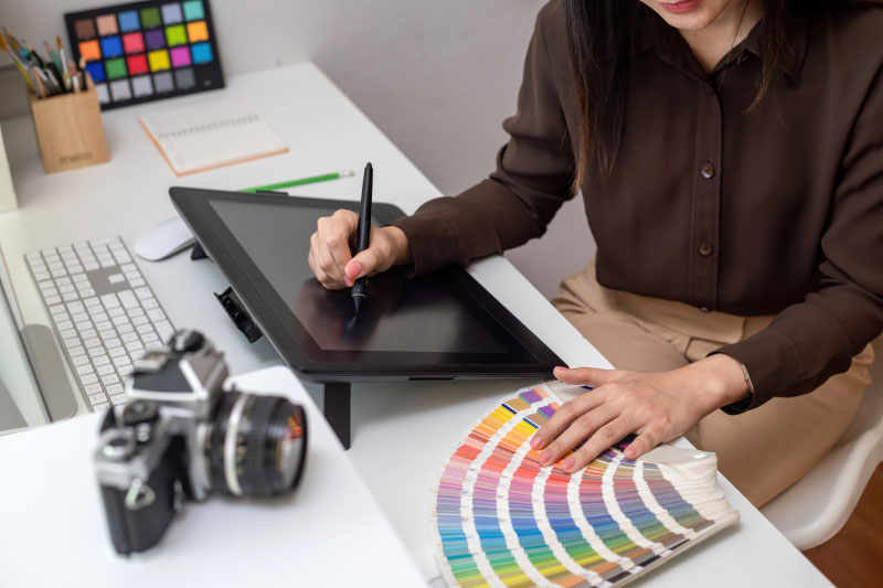 Grafik pracujący na tablecie graficznym nad projektem, obok leży wzornik kolorów i aparat, ilustrując proces tworzenia spójnej identyfikacji wizualnej i doboru kolorystyki marki.