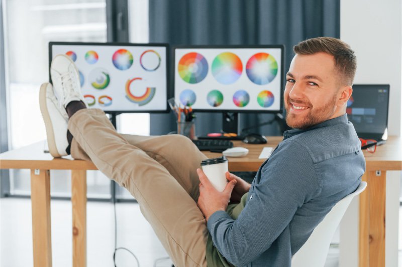 Zrelaksowany projektant stron internetowych przy dwóch monitorach pokazujących koła kolorów, ilustrujący łatwość i szybkość tworzenia witryn no-code we Framer bez potrzeby zatrudniania programisty.